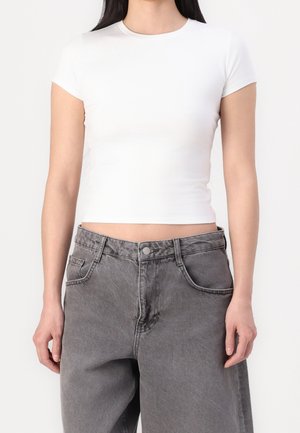Vrouw draagt een aansluitend wit cropped T-shirt en losse grijze jeans met hoge taille, staande tegen een effen achtergrond.