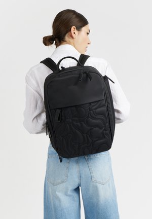 Donna che indossa una camicia bianca e jeans azzurri chiari porta uno zaino nero con parte frontale testurizzata e molte tasche con cerniera.