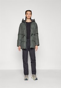 Giacca imbottita verde scuro con cappuccio, vita regolabile e design trapuntato. Abbinata a pantaloni neri e sneakers grigio chiaro.