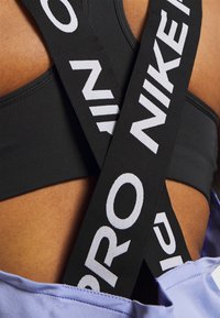 Czarny stanik sportowy z kontrastującymi białymi paskami logo Nike krzyżującymi się na lawendowej bluzce, podkreślający gładką fakturę i sportowy design.
