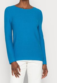 Maglione a maniche lunghe in teal a coste, con scollatura ampia e taglio dritto. Indossato su pantaloni bianchi, caratterizzati da una texture liscia e un design confortevole.