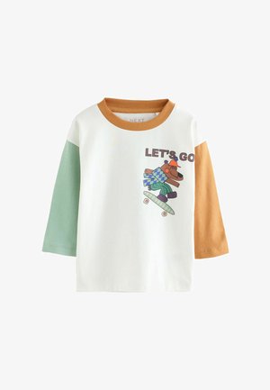 Camiseta blanca con mangas verdes y marrones, que presenta un gráfico de un oso haciendo skate y el texto "VAMOS" en marrón.