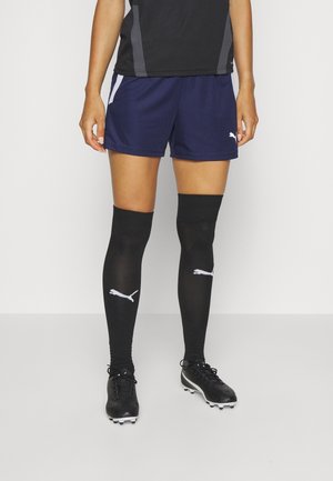 Personen har på med marineblå sportsshorts, sorte knæstrømper fra Puma og sorte fodboldstøvler, står mod en ensfarvet baggrund.