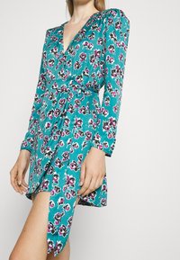 Tüll langärmliges Kleid in Türkis mit einem Blumenmuster in Pink und Schwarz, mit einem tiefen V-Ausschnitt und seitlichem Wickeldetail.