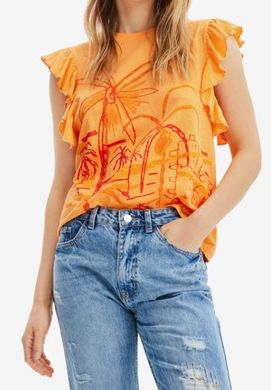 Kobieta ubrana w pomarańczową bluzkę bez rękawów z falbankami na ramionach i abstrakcyjnym czerwono-pomarańczowym wzorem, zestawioną z jasnoniebieskimi podniszczonymi dżinsami.