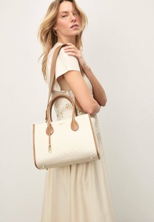 Woman in a beige dress holding a cream and tan handbag with double handles and a shoulder strap against a neutral background.