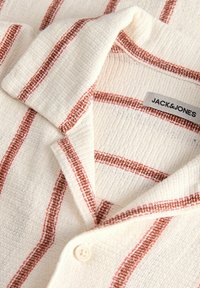 Chemise crème texturée avec des rayures verticales et horizontales couleur rouille, avec un col plié et un seul bouton visible.