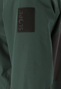 Veste étanche verte dotée d'un patch noir texturé avec l'inscription "SLOPE STYLE" sur le bras. Tissu lisse au design contemporain.