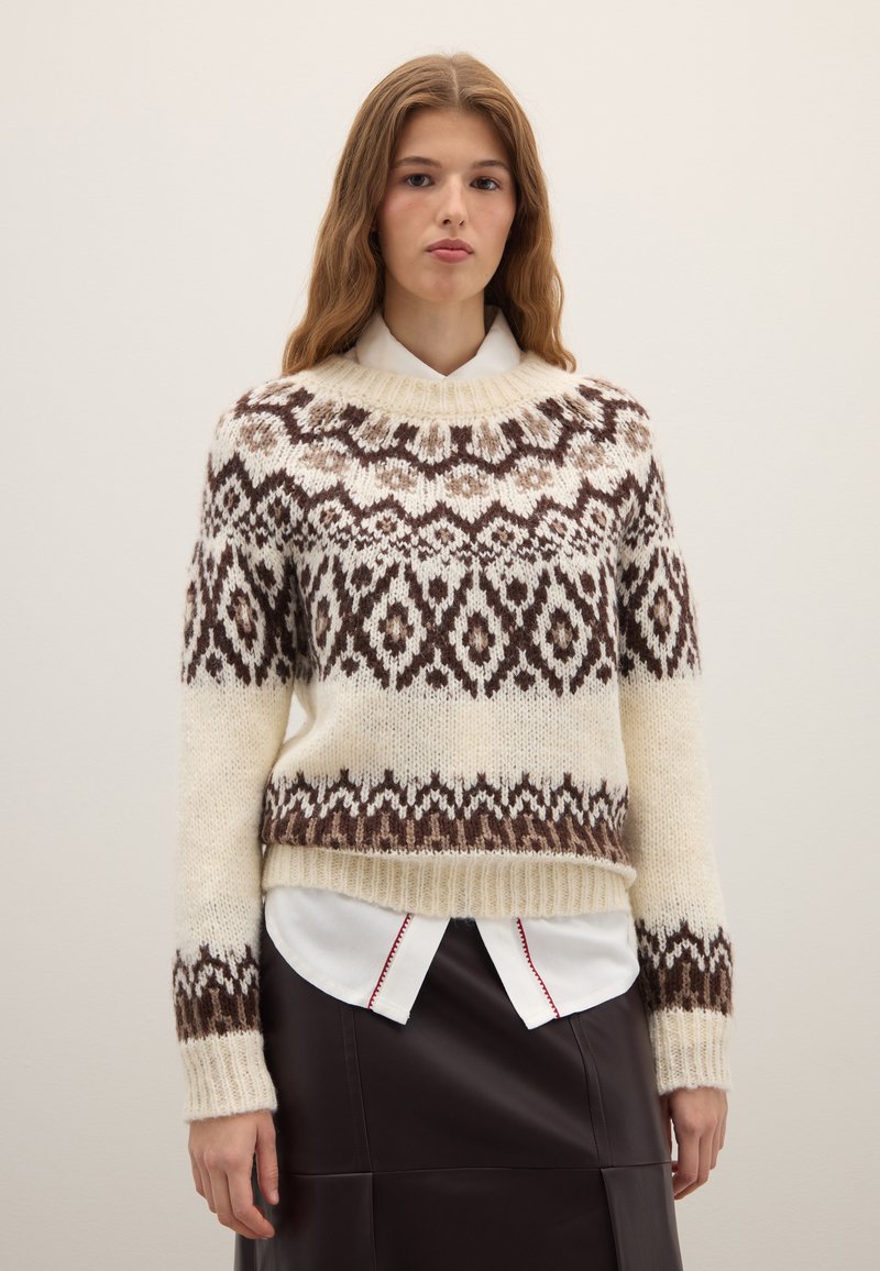 Dikgebreide trui in crèmekleur met bruin en beige geometrisch patroon, brede geribbelde zoom en boorden, ronde halslijn, gedragen over een wit overhemd.