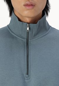 Pullover light blue a metà zip realizzato in morbido tessuto testurizzato. Presenta un colletto alto e una cerniera metallica al collo. Design minimalista.