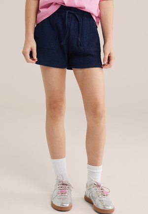 Person iført marineblå shorts med snøre, lyserød skjorte, hvide ribstrikkede crew-sokker og sølvfarvede sneakers med brune såler, stående mod en neutral baggrund.