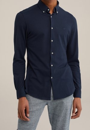 Man draagt een aansluitend donkerblauw, langarmig overhemd met knopen en lichtgrijze broek, staat met ontspannen handen langs het lichaam.