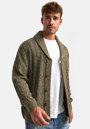 Mann, der einen grünen, kabelgestrickten Cardigan mit Knöpfen über einem weißen T-Shirt und blauen, abgetragenen Jeans trägt, steht vor einem weißen Hintergrund.