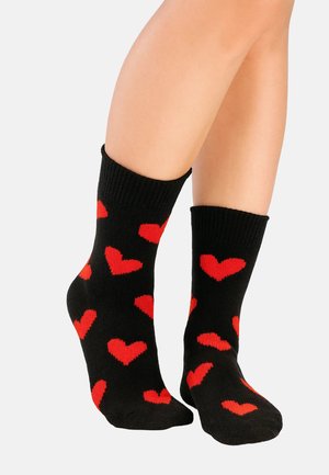 Schwarze Socken mit einem Muster aus roten Herzen. Sie sind aus einem weichen, dehnbaren Material mit einem gerippten Bündchen gefertigt und sitzen gerade über dem Knöchel.