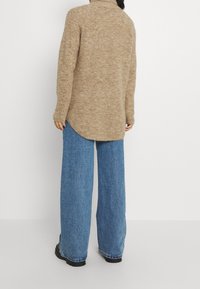 Suéter de punto beige con cuello alto, combinado con unos vaqueros de denim azul de corte ancho. El suéter tiene un dobladillo redondeado y mangas largas.