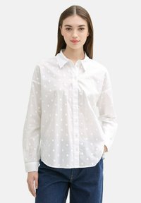 Weißes Hemd mit Knopfverschluss, gepunktet, lange Ärmel und Kragen. Hergestellt aus leichtem Stoff mit weicher Textur. Kombiniert mit blauen Jeans.