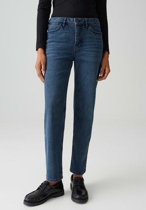 Jeans bleu foncé à coupe droite, dotés d'une fermeture à bouton, de poches avant et de coutures apparentes le long des coutures. Portés avec des chaussures noires.