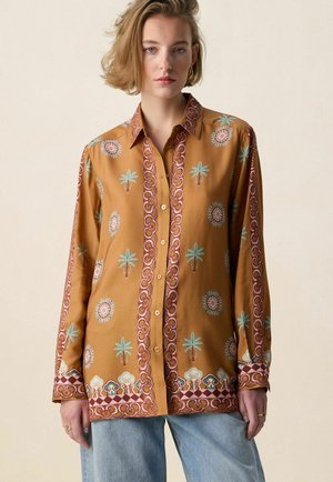 Femme portant une chemise boutonnée jaune moutarde avec des motifs de palmiers et des motifs circulaires, associée à un jean bleu clair, debout devant un fond uni.