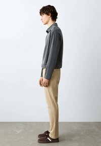 Tânăr în picioare, văzut din profil, purtând o cămașă gri cu mâneci lungi, pantaloni bej și adidași maro pe un fundal simplu, deschis la culoare.