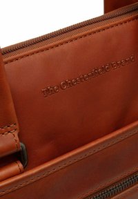 Sac en cuir brun avec une texture lisse, doté d'une fermeture éclair, d'un logo en relief et de détails de couture le long des bords.