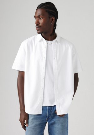 Camicetta bianca a maniche corte con colletto, realizzata in cotone, abbinata a una maglietta bianca e jeans azzurri chiari. Texture liscia, design minimale.