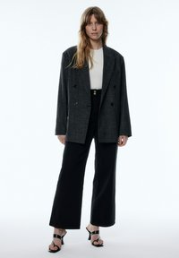 Grijze, dubbelzijdige blazer met zwarte knopen, gecombineerd met een witte top en zwarte, hoge wijdvallende pants. Zwarte, gespen sandalen.