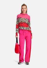 Rosa Pullover mit Leopardenmusterstreifen, kombiniert mit knallpinkenSweatpants und einer roten Tragetasche. Die lässigen Schuhe haben ein Leopardenmuster.