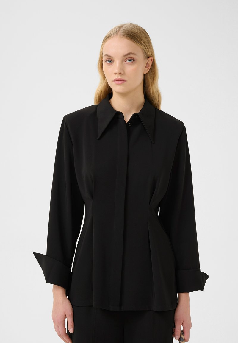 Blusa nera a maniche lunghe con colletto strutturato, design frontale plissettato e polsini leggermente svasati, realizzata in tessuto morbido.
