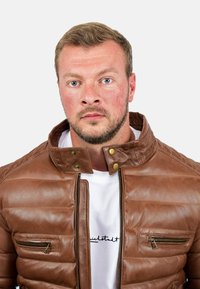 Giacca di pelle marrone con design imbottito, colletto alto e dettagli in metallo dorato. Presenta due tasche con zip e una maglietta bianca sotto.