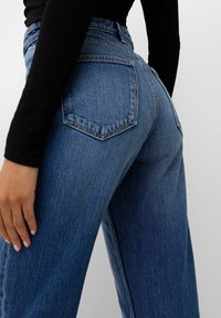 Modré džíny s vysokým pasem, zadními kapsami, rovnými nohavicemi a texturovanou tkaninou. Viditelné stehy a mírné vyblednutí na bocích.