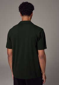 Polo shirt verde scuro con una vestibilità rilassata, maniche corte e colletto classico. Tessuto liscio, senza motivi visibili o dettagli in metallo.