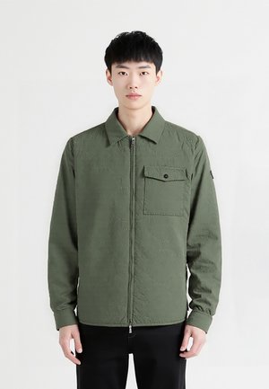 Olijfgroene zip-up jas met een kraaghals, een voorzak, gemêleerde stof en lange mouwen, gecombineerd met zwarte broek.