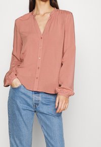 Blouse rose avec manches longues, col rond, accents froncés et fermeture à boutons. Associée à un jean en denim bleu clair.