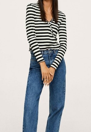 Femme portant un haut rayé noir et blanc à manches longues et un jean bleu taille haute, debout les mains jointes devant.