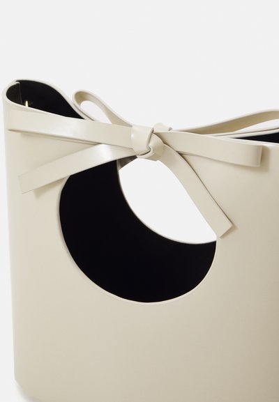 Cream-colored handbag in a semi-circular shape, featuring a cutout handle, tied ribbon detail, and a contrasting black interior.