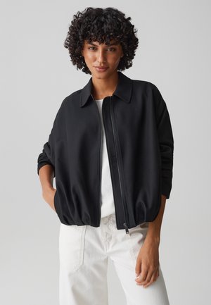 Frau mit lockigem Haar, die eine schwarze Jacke mit Reißverschluss über einem weißen Oberteil und weißen Hosen trägt, steht mit einer Hand in der Tasche.