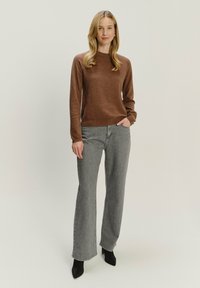 Vrouw die met één hand in de zak staat, gekleed in een bruine lange-sleeve trui, grijze rechte jeans en zwarte puntige laarzen op een effen achtergrond.