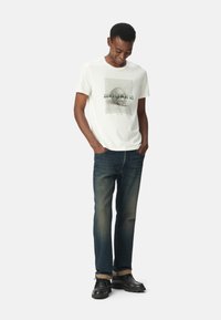 Camiseta blanca de algodón con un gráfico de calavera y el texto "NO NOS MIRES", combinada con unos pantalones vaqueros oscuros y calzado negro.