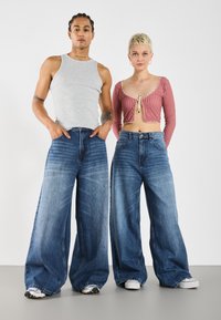 Weite blaue Jeans aus Denim mit heller Waschung, hochgeschnitten und mit subtilen Verwaschungen. Kombiniert mit einem grauen Tanktop und einem roten Karo-Crop-Top.