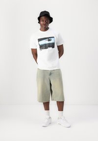 Camiseta blanca con un estampado gráfico; pantalones cortos de mezclilla claros; sombrero tipo bucket negro; zapatillas blancas; el modelo está de pie contra un fondo blanco liso.