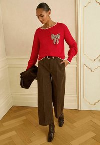 Roter Pullover mit einem Leopardenmuster und Schleifendetail, mit gewelltem Ausschnitt. Braune weit geschnittene Hose, kombiniert mit einer schwarzen Tasche und braunen Stiefeletten mit Absatz.