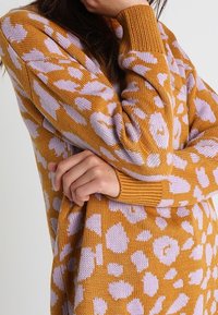 Garnattröja i senapsgul med stora lila fläckar. Har ribbade muddarna och en avslappnad passform som visar en texturerad mönster.