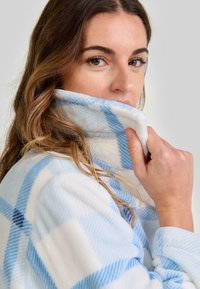 Mujer con cabello ondulado sube el cuello de una suave chaqueta de forro polar a cuadros blancos y azules, mirando por encima de su hombro.