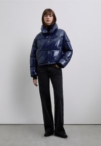 Donkerblauwe pufferjas met een glanzende afwerking, hoge kraag en knoopsluiting, gecombineerd met zwarte uitlopende broek op een lichtgrijze achtergrond.
