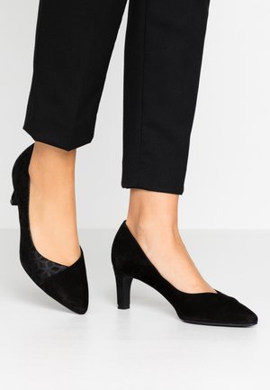 Kobieta w czarnych zamszowych butach na obcasie typu kitten heel, zestawionych z czarnymi spodniami 3/4 na białym tle.
