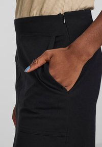 Pantalon noir au design ajusté, doté d'une poche latérale et d'une fermeture éclair à la taille. Texture lisse et style minimaliste.
