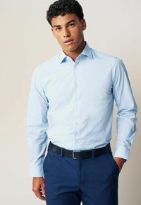 Chemise à manches longues bleu clair, texture lisse, avec un col classique et une coupe ajustée. Portée avec un pantalon bleu marine et une ceinture noire texturée.
