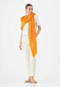Écharpe légère orange drapée sur un t-shirt blanc, accompagnée de jeans clairs et de sandales tissées beige, le tout placé sur un fond blanc.