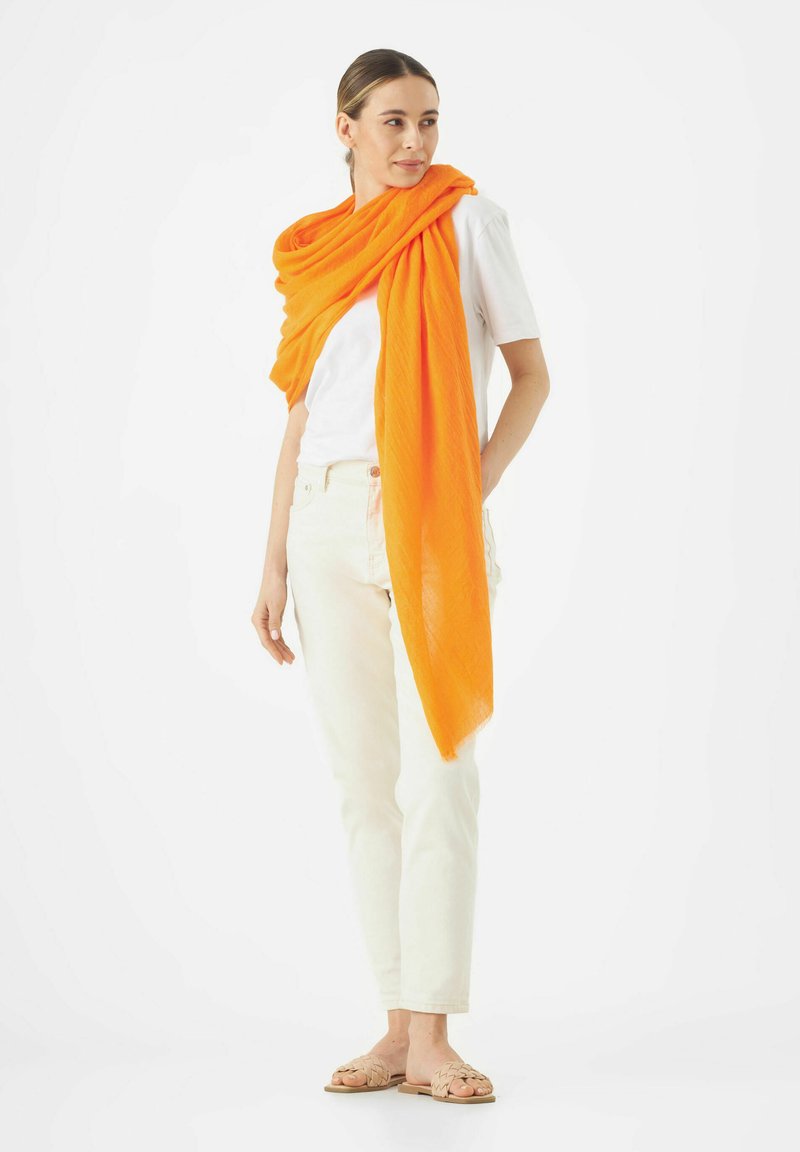 Écharpe légère orange drapée sur un t-shirt blanc, accompagnée de jeans clairs et de sandales tissées beige, le tout placé sur un fond blanc.