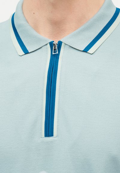 Polo bleu clair avec un col rayé gris et bleu et une fermeture éclair centrée rayée bleu et blanc, partiellement zippée au niveau du col.